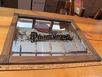 Full front view of the Pilsner Urquell mirror showing the wooden frame and etched glass with logo and emblem, placed on wooden table with measuring ruler adjacent showing width.