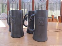 Four matte grey ceramic beer mugs arranged on a wooden table showing the embossed Playboy bunny logos on their fronts.
