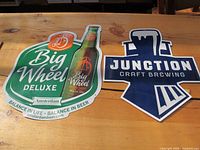 Front view of two tin beer signs: Big Wheel Deluxe Amsterdam and Junction Craft Brewing on wooden table