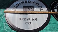 Photo of round tin sign with 'Refined Fool Brewing Co.' text and a ruler showing diameter measurement around 23.5 inches.