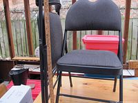 Side and front views of a black metal folding chair with cushioned seat and back in striped fabric, showing approximate seat height with a yardstick.