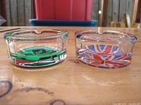 Photo showing two glass ashtrays side by side on a wooden surface. One ashtray has a green cannabis leaf design, and the other has a British Union Jack flag design. Both have three cigarette notches.