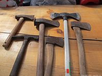 Overview of five axes and one rock hammer laid on a wooden surface, showing handles and blades.