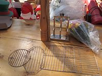 Photo showing three metal cooking grates of different shapes laid flat on a wooden table, a stainless steel cutlery holder, and an artificial flower bouquet wrapped in clear plastic.