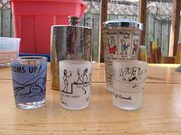 Front view of shot glasses, metal hip flask, and stacked shot glasses with colorful designs on wooden table