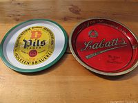 Two metal beer trays on wooden surface: red Labatt's Ale tray with gold rim and yellow, white and green Holsten-Brauerei Pils Lager tray