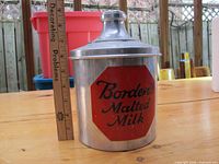 Front view of the vintage Borden's Malted Milk metal tin container with lid, placed next to a ruler for size reference on a wooden surface outdoors.