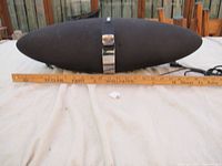 Elliptical black speaker with fabric covering and chrome band with logo, placed on a table alongside a 12-inch ruler for scale showing approximately 23 inches width.