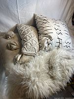 Four decorative pillows arranged on a surface alongside a white faux fur throw. Visible pillows include two white square pillows with different black stitched patterns, a rectangular cream macrame knit pillow with pom-poms, and the fluffy white faux fur throw.