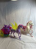 Photo showing the purple horse with yellow butterfly wings attached to a colorful butterfly-themed carriage with rolling wheels, next to a white horse with pink mane and pink saddle.