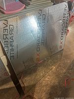 Large glass mirror with reflective surface showing protective foil on one side and concrete floor beneath.