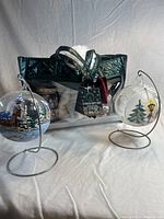 Clear tote bag containing the gift set items alongside two glass sippers with painted holiday glass ornaments, placed on metal stands.