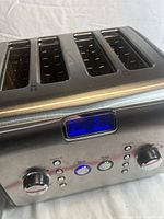 Close-up front angle showing four slots, digital blue display indicating toast levels, and control buttons/knobs for browning, toast, and warm.