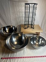 Full lot showing three stainless steel mixing bowls, black metal wire storage rack on wooden footstool with measuring tape for scale