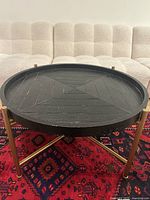 Full view of the round black wood coffee table with brass legs placed on a red patterned rug.