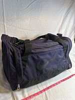 Side view of purple Nike gym bag showing main compartment, black handles, and adjustable shoulder strap.