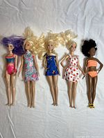 Five Barbie dolls lying on white fabric background, each with a single distinct outfit and varied hair colors.