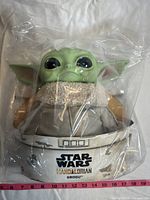 Front view of Grogu toy with large black eyes, green skin, and gray robe, in packaging with measuring tape indicating size.