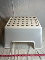 Front view of white plastic step stool showing black circular grips on the top surface and overall shape and size with tape measure.