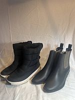 Front and side view of two pairs of women's boots placed next to each other on a white backdrop. One pair is black insulated winter boots with white soles, the other is black Chelsea boots.