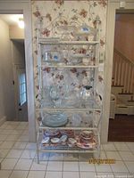 Full view of the wrought iron baker's rack with multiple glass shelves holding glassware in front of floral wallpaper background.