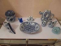 Overview of blue and white porcelain pieces, blue glass items including cat shakers, rooster creamers, dragon teapot, Victorian shoe, and blueglass wine decanter with cap displayed on white table.