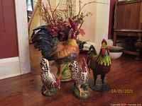 Four rooster-themed items arranged on a wooden floor: large ceramic planter, metal figurine, two smaller ceramic roosters.