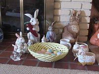 Group photo of all rabbit figurines along with yellow lattice basket and decorative eggs placed on a brick surface in front of a fireplace.