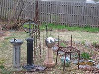 Overall view of garden items including sundial on pedestal, spiral willow obelisk, metal plant trellis, stone columns, metal rooster, wrought iron chair, and decorative globes arranged on grass.