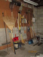 Wide view of multiple garden tools hanging on pegboard and leaning against wall in garage space, showing wooden-handled rakes, shovels, and other tools.