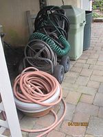 View showing multiple garden hoses coiled on and off a metal wheeled hose cart alongside two plastic outdoor trash cans.