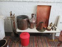 Full view of vintage copper bucket, watering can, antique pitcher, copper tray, six brass bells, paperweight, brass fireplace tools, and red metal bucket arranged on and beside a white bench.