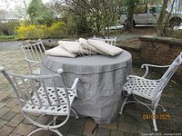 Photo of round metal table covered with a fitted protective cover and four lattice design metal arm chairs surrounding it with four seat cushions on top.