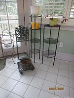 Four metal plant stands of varying heights with flower pots and a vintage cast iron trivet on tiled floor near windows.