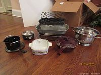 Full lot showing all cookware items arranged on floor including stoneware pot, colander, bake dish, sauce pot, roasting pans, and toast racks.