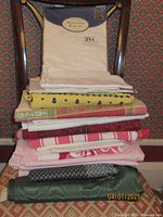 Stack of folded assorted table covers in various patterns and colors, including light pink, red striped, green textured, a yellow floral patterned cover, and a packaged white Heirloom Eyelet tablecloth on top.
