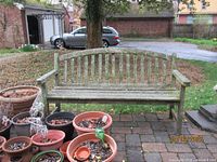 Front view of the teak wood bench showing weathering and moss growth with surrounding flower pots and brick patio.