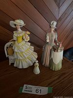 Two larger Avon porcelain figurines depicting Victorian women and one smaller figurine on wooden surface with angled wood wall background