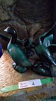 Two green ceramic geese figurines, one in flight with wings raised and one standing, shown on a stone surface.