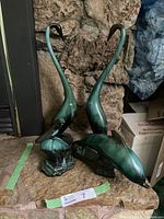 Two tall ceramic long neck birds and a dolphin shown together on a stone surface in front of a stone wall.