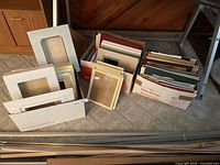 Three cardboard boxes filled with assorted cut mats and shadow boxes placed on carpet near wood-paneled wall.