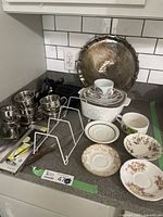 Photo showing six metal coffee cups with saucers, silver-plated tray, Corning casserole with lid, and several vintage teacup saucers on a kitchen counter.