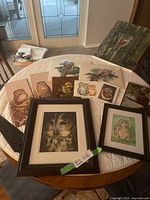 Photo showing multiple owl artwork pieces arranged on a round table, including framed and unframed prints depicting different owl species