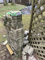 Tall stack of about 50 concrete paving stones on grass next to wooden lattice fence, with some loose stones around it
