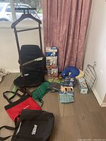 Photo showing a collection of personal care and support items arranged near a window and curtain, including a wooden valet butler, black massaging seat cushion, blue neck pillow, boxed fans, grab bar, tote bags, and a green bag.