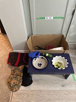Full view of the lot showing the bowl stand with two bowls on top, colorful dog jacket, leashes, green portable water bottle, and cardboard box with other essentials.
