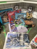 Lot visibly includes assorted animal themed boxed jigsaw puzzles, partially completed puzzles and a wolf diamond paint canvas, displayed on a green surface against white subway tile.