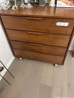 Front view of the teak dresser showing four drawers and wood finish, with small wheels at the base.
