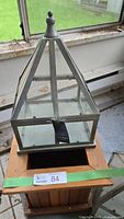 Metal and glass pyramid terrarium placed on top of wooden planter box, showing small terrarium with pyramid top and finial knob, with visible window and siding in background.