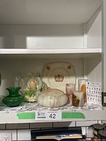 Shelf display showing stone bowl, candle holders, ceramic plate, green vase, glass dome bird figurine, and small card